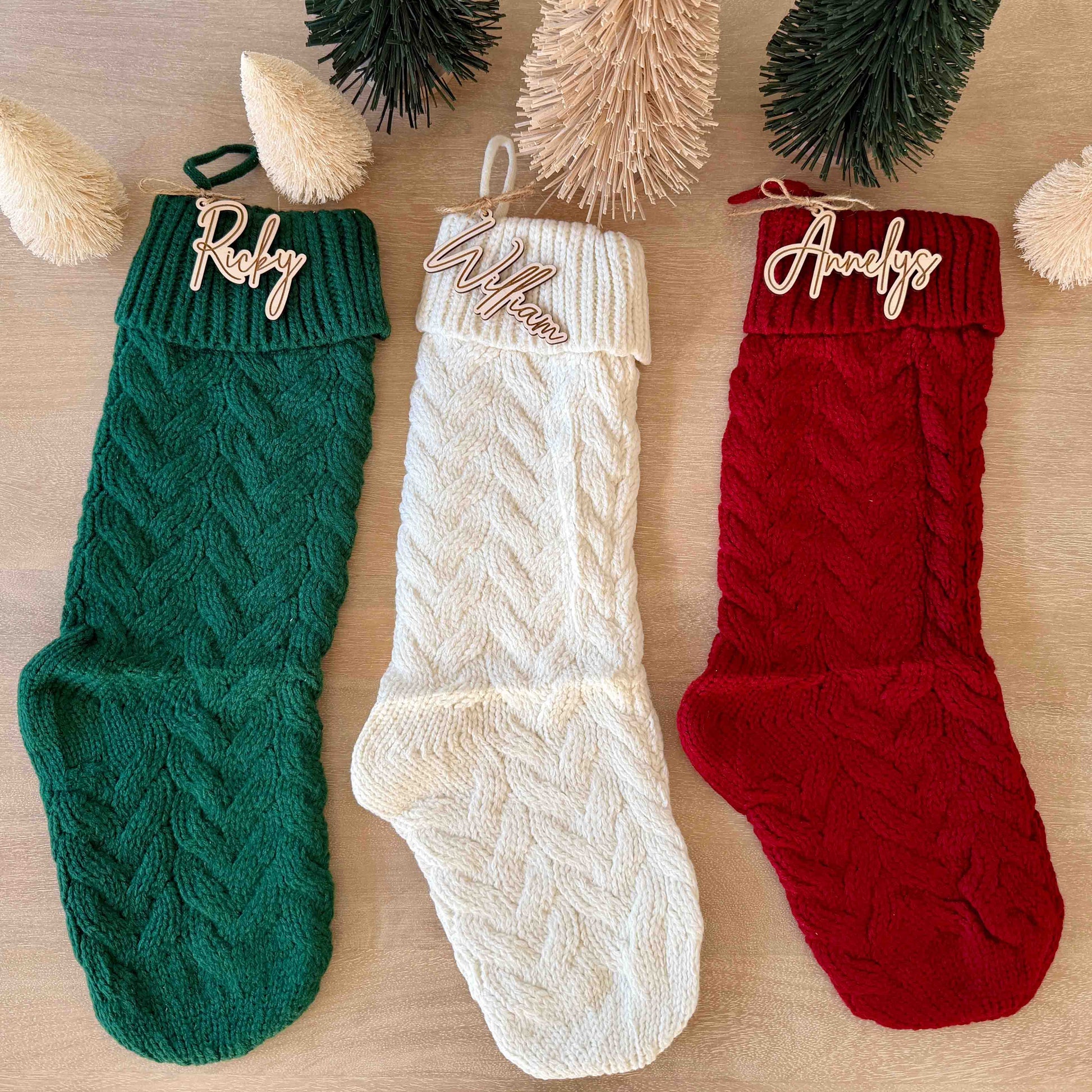Set of three knit Christmas stockings in green, white, and red, each accented with a personalized wooden script name tag.