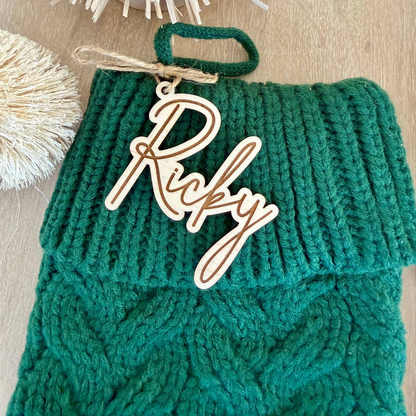 Green knit Christmas stocking with engraved wooden script name tag reading “Ricky,” tied with rustic twine.