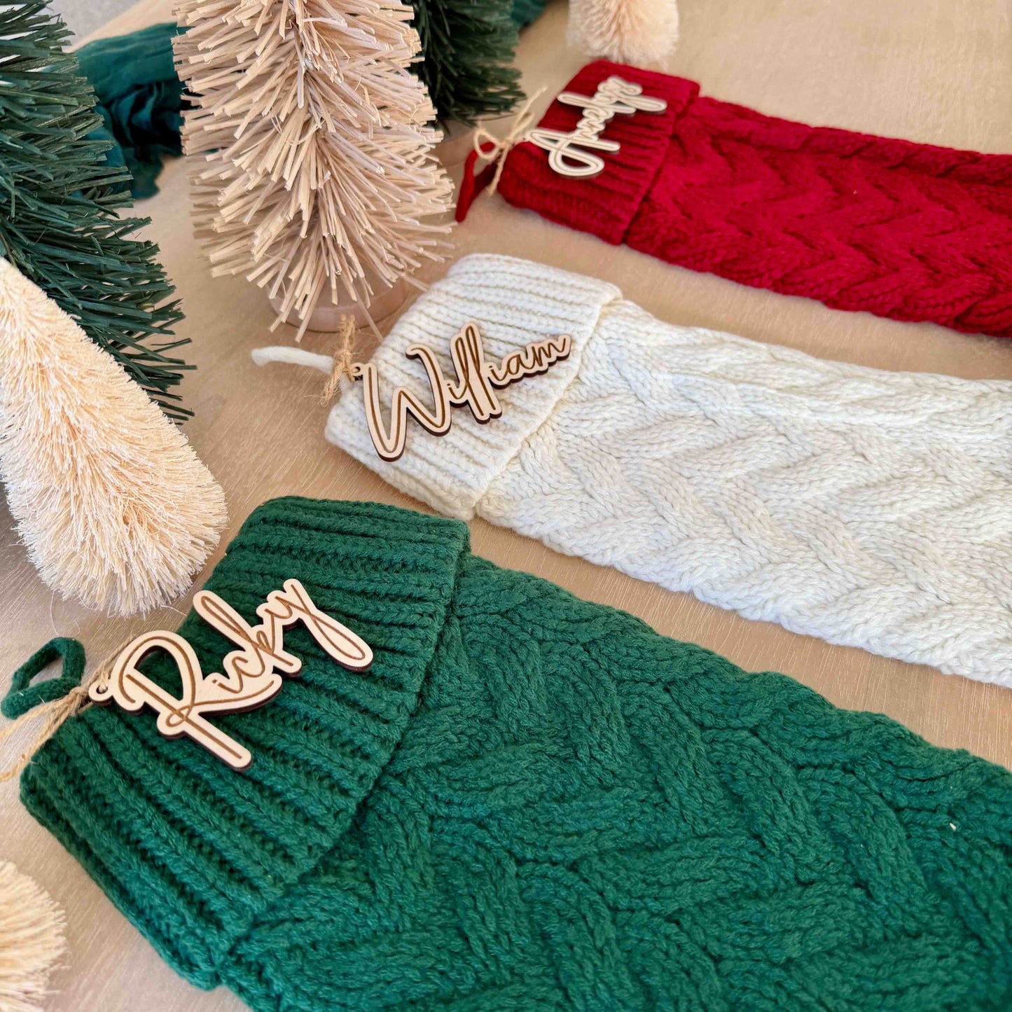 Overhead view of knit Christmas stockings in three colors, each with a custom wooden name tag tied on with twine.